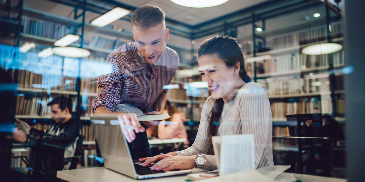 A librarian and graduate student collaborate over a laptop in a library, surrounded by bookshelves.